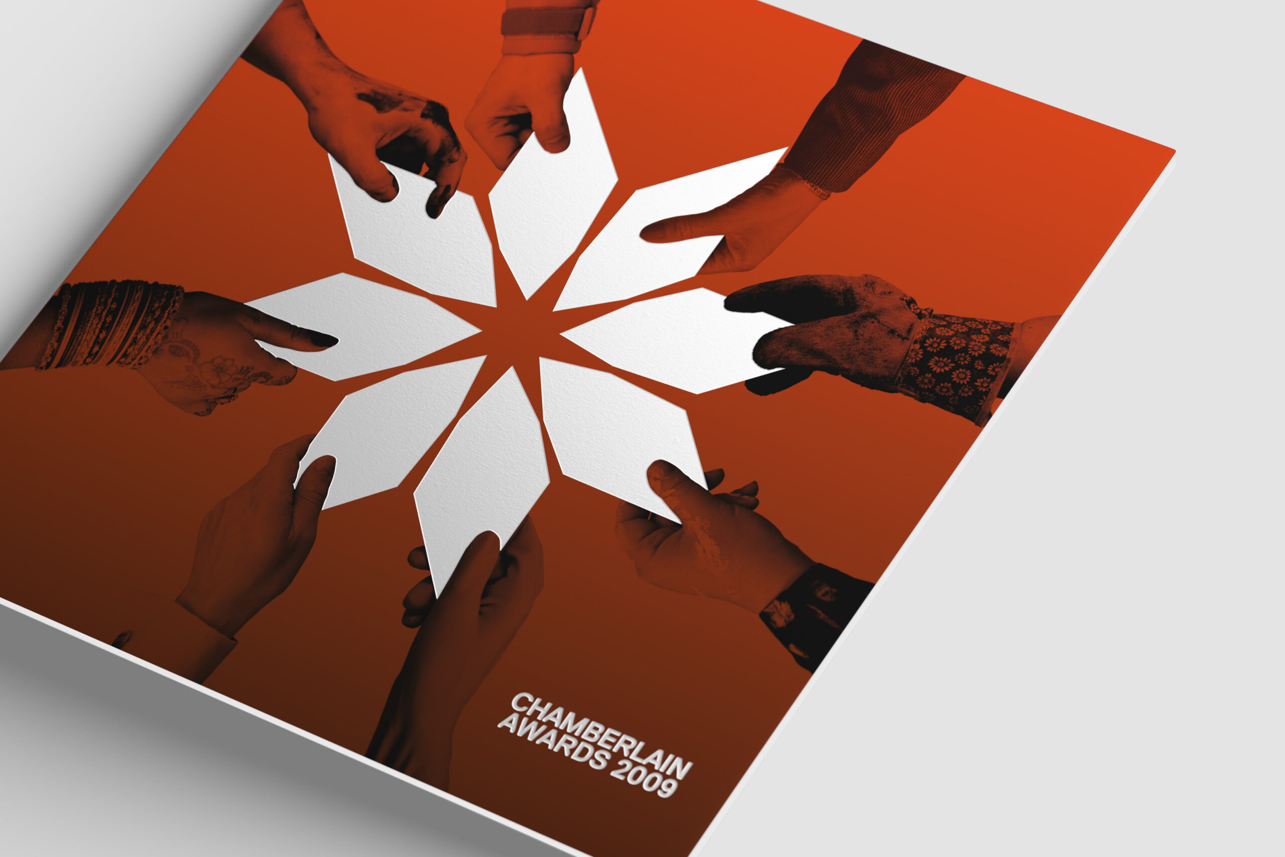 The Chamberlain Awards 2009 brochure sits on a neutral grey background. The cover image is printed in shades of orange on a heavy uncoated board. The star-shaped logo sits at the centre, embossed in white. The logo is made of of diamond-shaped lozenges held by people in a variety of clothing and gloves.