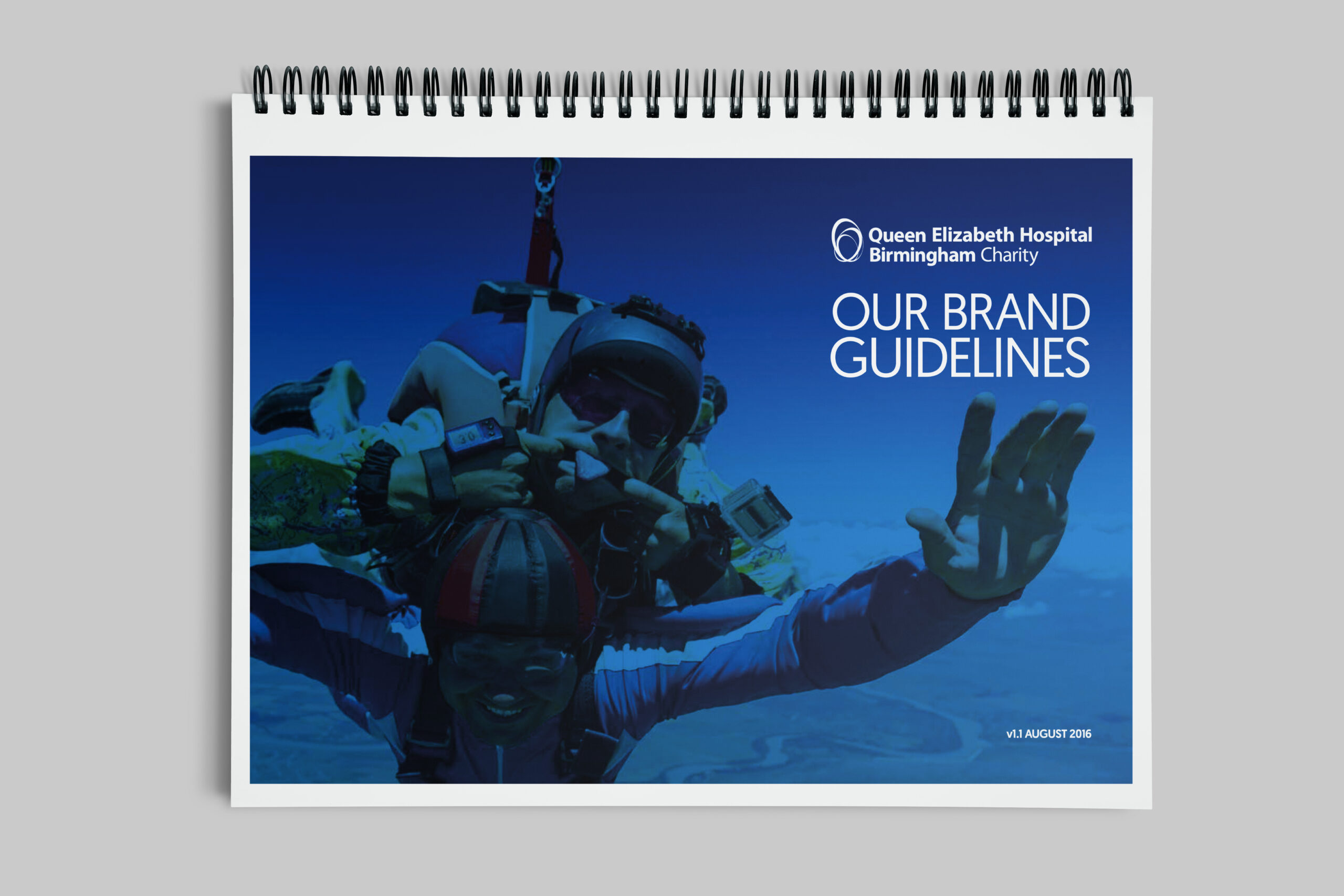 Image of the wire-bound QEHB Charity Brand Guidelines edition 1. The cover shows a comical image of a tandem skydive with the participants smiling and pulling funny faces