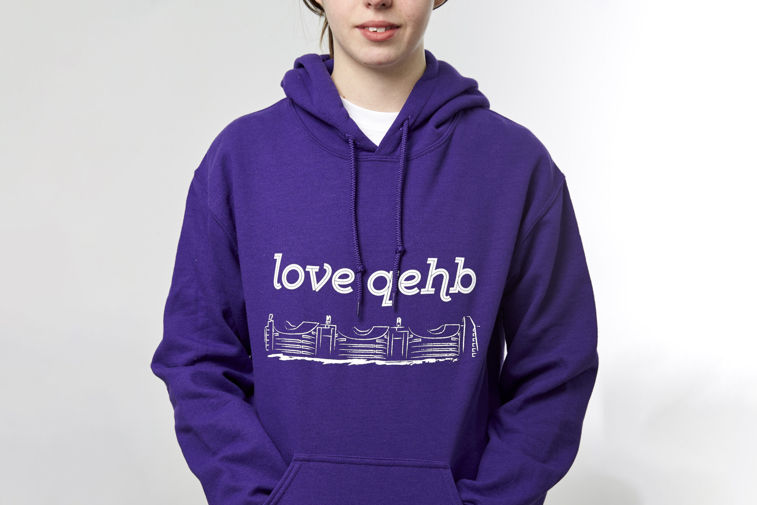 A smiling model wears a purple hoodie incorporating a hand-drawn image of the Queen Elizabeth Hospital Birmingham. Above the drawing are the words 'Love QEHB'