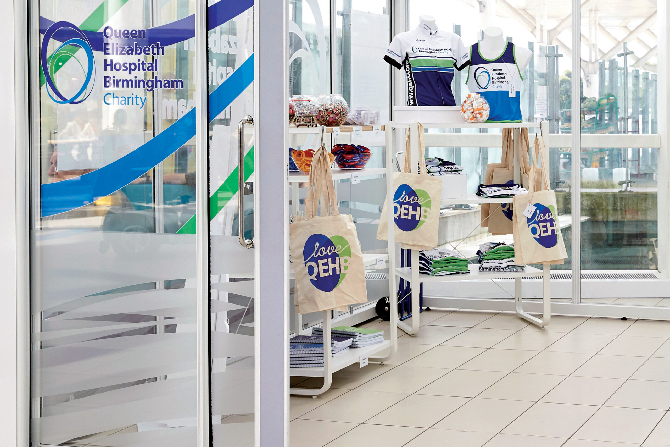 Image of the QEHB Charity retail kiosk in the foyer the Queen Elizabeth Hospital Birmingham. Inside the glass-walled shop are shelves containing QEHB branded clothing, stationery and accessories.