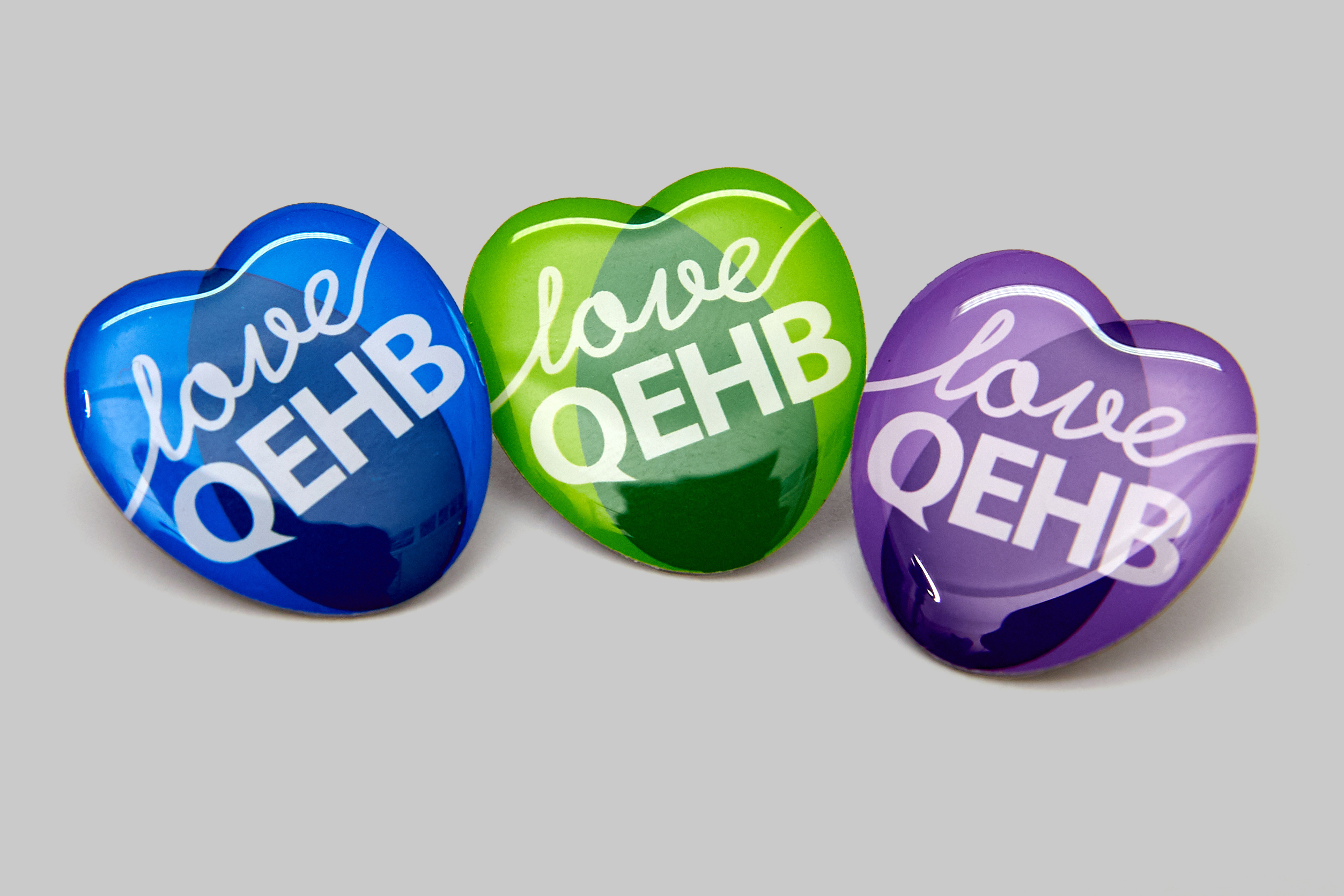 Three heart-shaped pin badges are arranged on a neutral grey background. The words 'Love QEHB' are overlaid in white on the blue-, purple- and green-coloured badges.