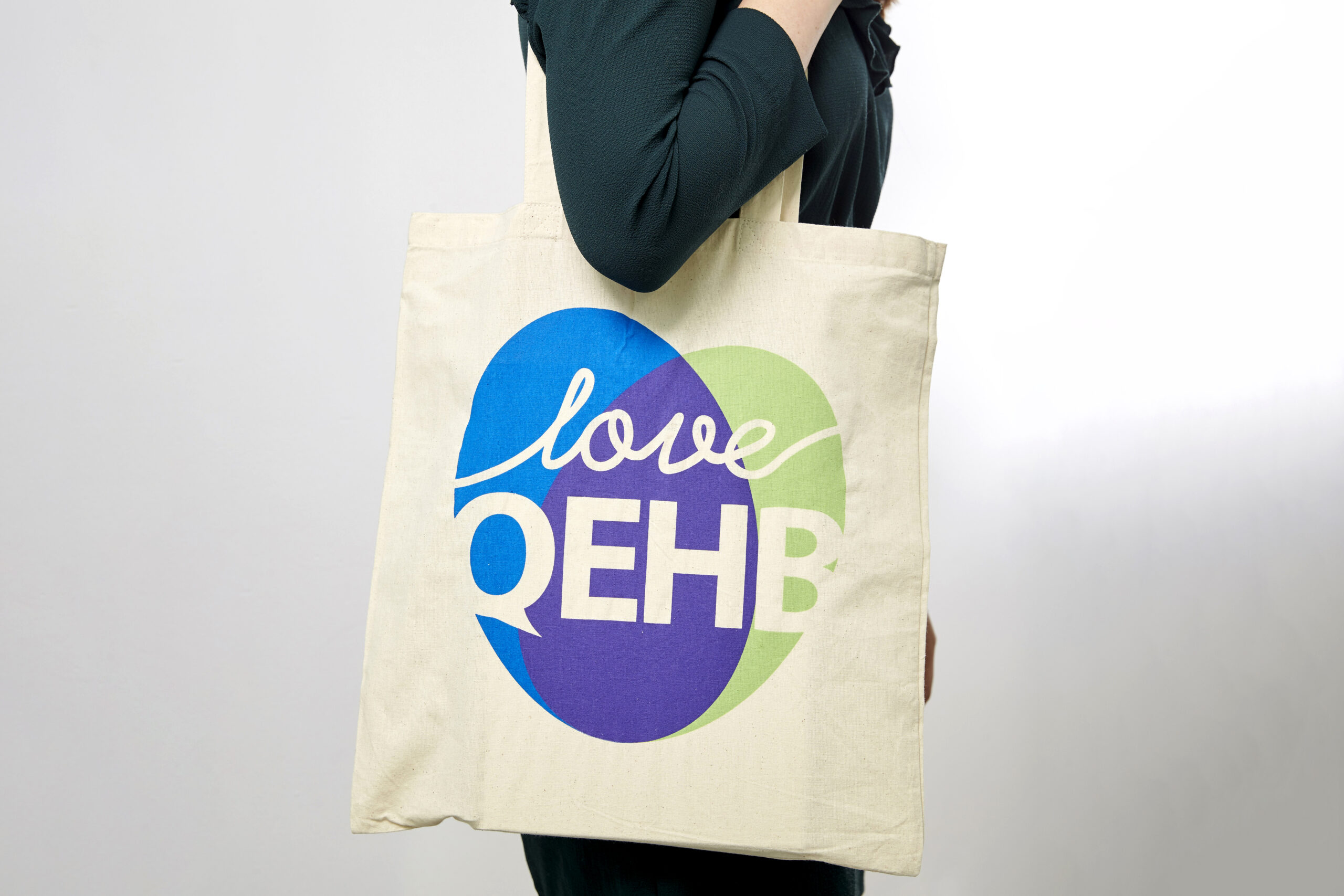 A model wears a QEHB Charity branded jute bag on her shoulder. The bag is printed with a green and blue overlapping ovals, forming a heart shape. Overlaid on the heart are the words 'Love QEHB'
