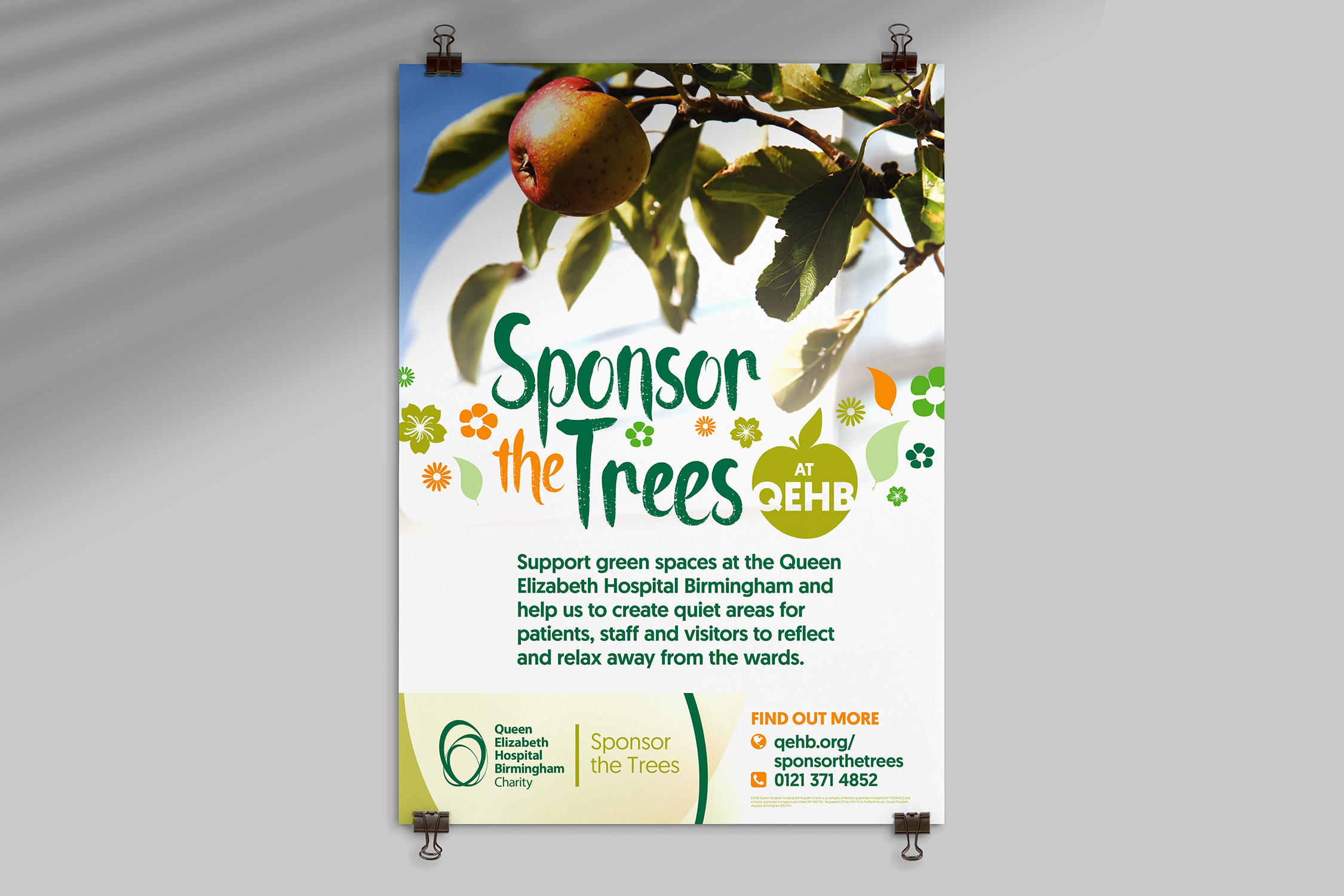 An A1 poster is clipped to a neutral grey wall. The poster shows a red apple hanging from a tree, with one of the Queen Elizabeth Hospital Birmingham's towers blurred in the background. Under the image the words 'Sponsor the trees at QEHB' are displayed in a playful handwritten font intermixed with images of flowers and fruit.
